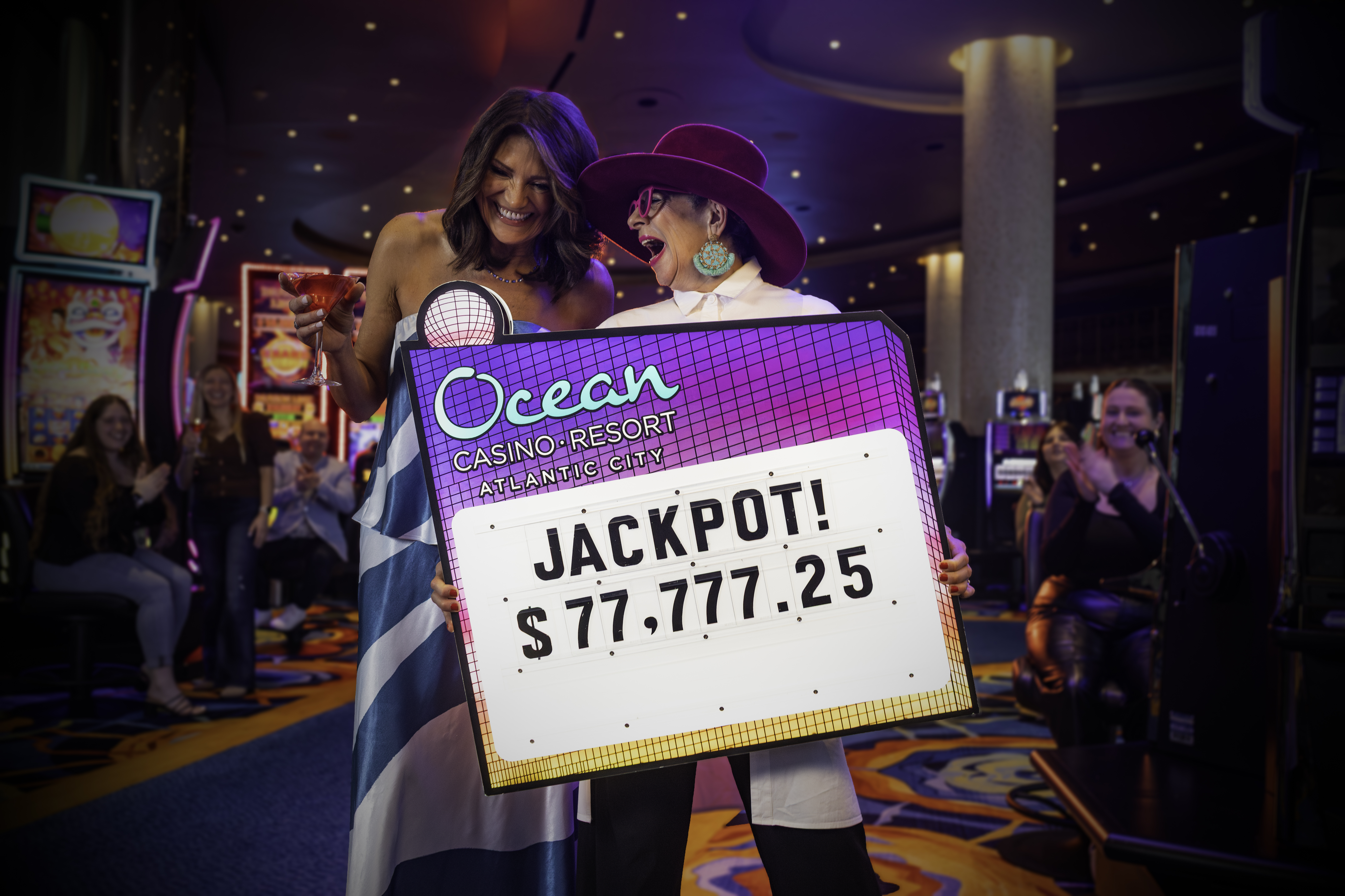 With cocktails in hand and wide smiles, two winners pose with a vibrant jackpot sign on the casino floor at Ocean Casino Resort as a crowd applauds behind them.