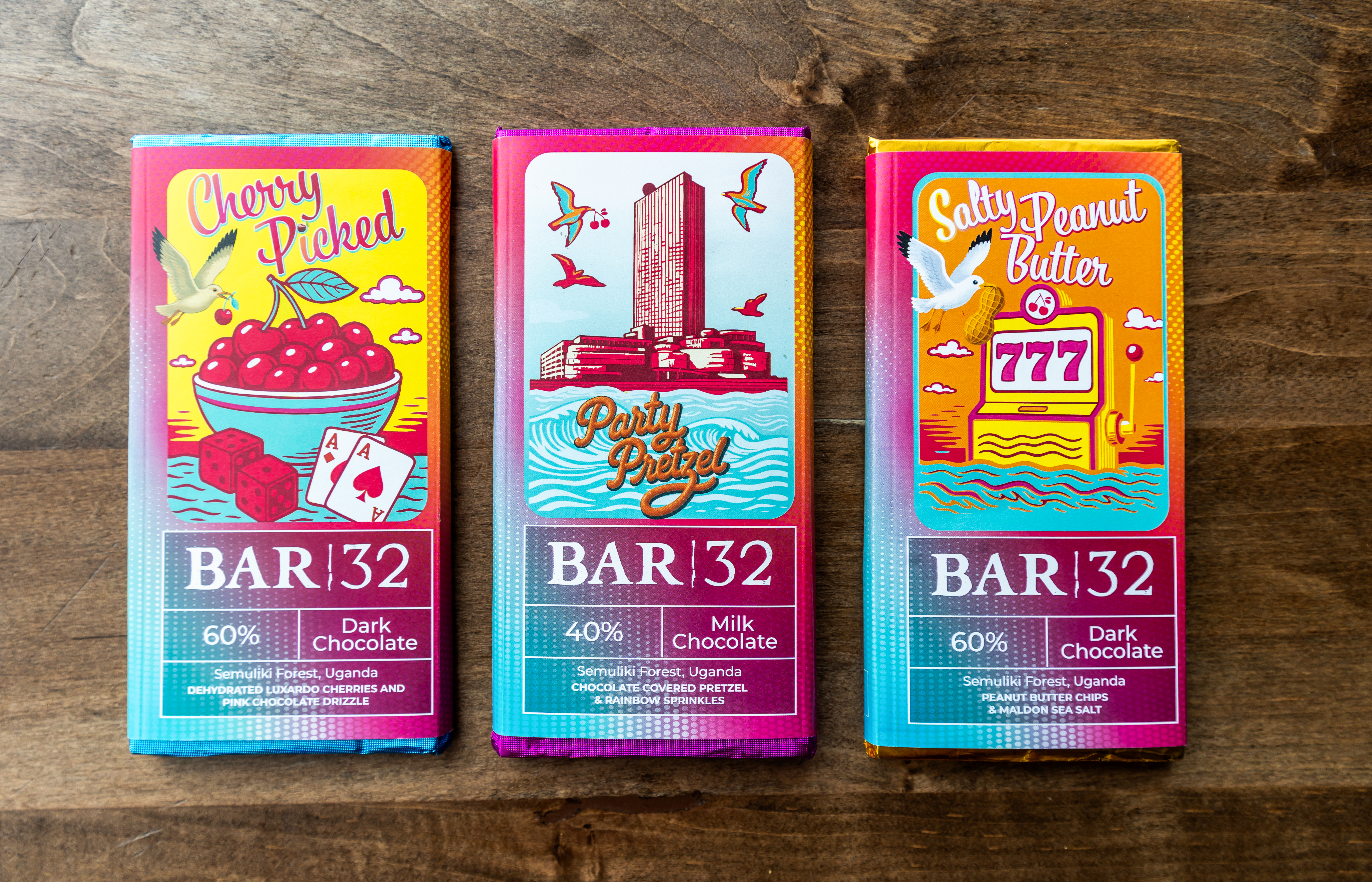 Three custom BAR 32 chocolate bars from Ocean Casino Resort displayed side-by-side on a wooden surface, each with vibrant packaging: "Cherry Picked" with cherries and playing cards, "Party Pretzel" featuring a stylized resort and seagulls, and "Salty Peanut Butter" with a slot machine showing 777.