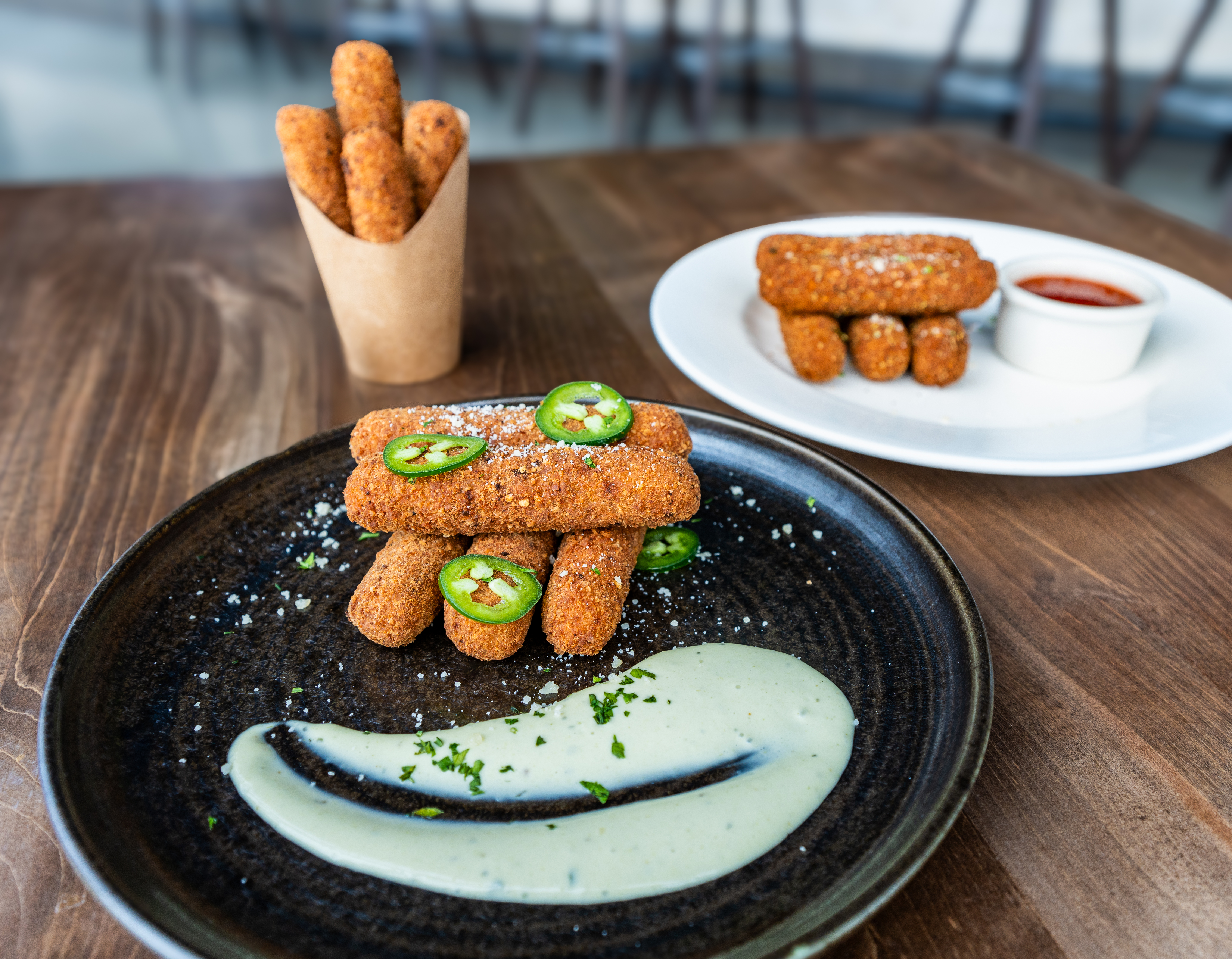 Close-up of crispy fried mozzarella sticks garnished with sliced jalapeños, served on a dark ceramic plate with a drizzle of creamy green dipping sauce, with additional mozzarella sticks and marinara sauce in the background.